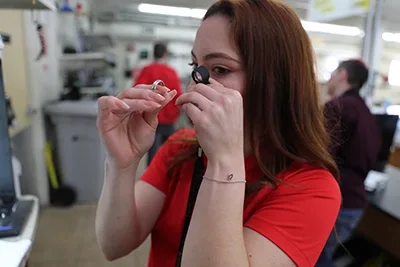 Woman in red shirt examining a diamond ring through a jeweler's loupe in a pawn shop.