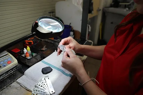 Woman in red shirt examining diamonds on a ring sizer at workbench with magnifying lamp and tools.