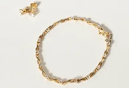 Gold bracelet with twisted design and embedded clear stones, paired with a set of matching stud earrings, displayed on a light surface.