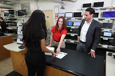 Sales associate in red shirt assisting two customers at electronics store counter with laptops and monitors displayed on shelves in background